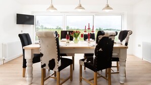 A dining area with a wooden table, four cowhide chairs, a vase with red flowers, and a view of a green field through large windows. - WESTWOOD COTTAGE - A spacious farm cottage close to superb East Lothian beaches  (Longniddry)