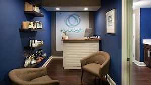 A reception area with a blue wall, a reception desk, two chairs, and shelves with various items.