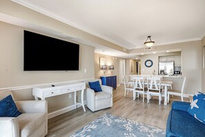 A modern living room with a flat-screen TV, white furniture, a dining area, and a kitchen in the background.