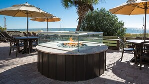 A circular outdoor seating area with a glass enclosure and a fire pit, surrounded by tables and chairs, with umbrellas and a palm tree in the background.