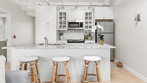 A modern kitchen with white cabinetry, a marble island, and three wicker barstools. - The Saltbox - Watch Hill's Award-Winning Waterfront Retreat.  (Westerly)