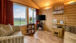 Ein gemütliches Blockhaus-Interieur mit Holzbalkendecke, einem großen Fenster mit Vorhängen, einem gestreiften Sofa, einem Bücherregal, einem Fernseher und einem kleinen Tisch.