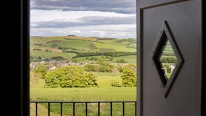 Blick durch eine Tür mit einem rautenförmigen Fenster auf eine grüne Landschaft mit sanften Hügeln und einem kleinen Dorf.