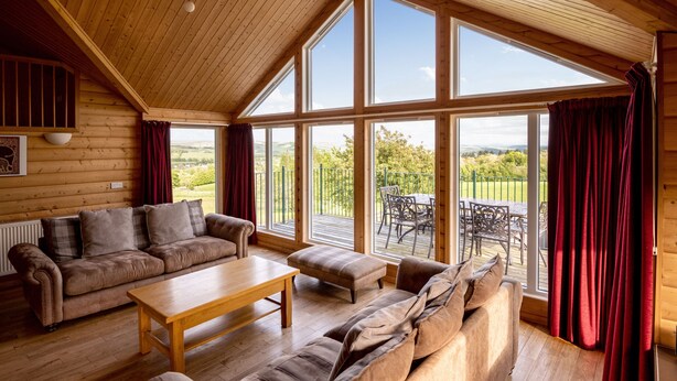 A log cabin interior with a wooden ceiling, large windows, a sofa, a table, and a view of the outdoors. - The Buchanan - sleeps 6 guests  in 3 bedrooms with hot tub - 5 star award winning lodges (Lauder)