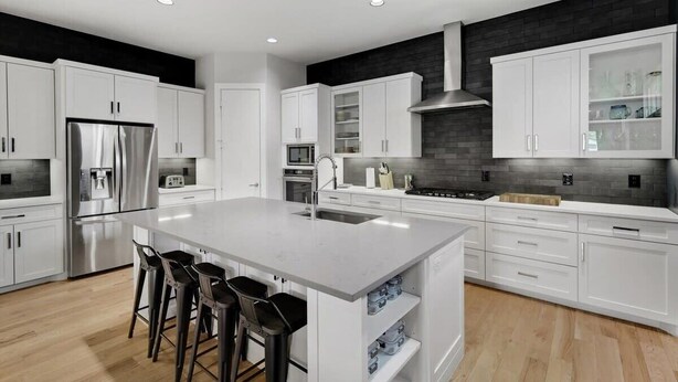 A modern kitchen with white cabinetry, a stainless steel refrigerator, a central island with bar stools, and a black brick backsplash. - Modern Luxury Townhouse | Uptown & Attractions (Dallas)