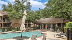 A pool area with lounge chairs and umbrellas, surrounded by trees and a building with a balcony. - Heart of Dallas - Cozy Apt, 2Bed, Pool (30+ days) (Dallas)
