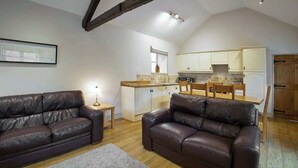 A cozy living room with two leather sofas, a small table, and a kitchen area with white cabinets and wooden flooring. - Host & Stay | Derwent (Consett)