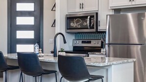 A modern kitchen with white cabinets, a stainless steel refrigerator, a microwave, and a built-in oven. There are three black bar stools and two pendant lights. - The Silo - Unique Experience, Lake, Firepit, BBQ (vian)