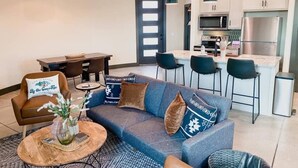 A modern living room with a sofa, coffee table, and chairs. A kitchen with a refrigerator, microwave, and dining area is visible in the background. - The Silo - Unique Experience, Lake, Firepit, BBQ (vian)