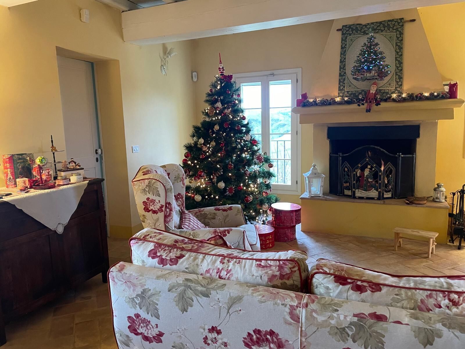 A living room with a Christmas tree, floral armchairs, a fireplace, and a mantel decorated with holiday ornaments.