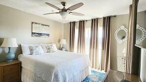 Une chambre à coucher avec un lit, deux tables de chevet avec des lampes, un ventilateur de plafond, un grand miroir et une fenêtre avec des rideaux.