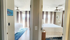 Une chambre d’hôtel avec deux lits, un ventilateur de plafond, une grande fenêtre avec des rideaux, un tapis bleu et une œuvre d’art murale.