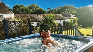 A hot tub with two people in it, surrounded by a wooden deck and a fence. - Pen Dragon - 2 Bed Cottage - Penclawdd (Swansea)