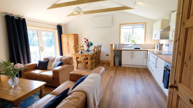 A spacious living area with a wooden dining table, leather sofas, and a kitchen with white cabinets and a wooden countertop. - Pen Dragon - 2 Bed Cottage - Penclawdd (Swansea)