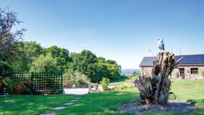A rustic stone building with a green fence, a tree stump sculpture, and a clear sky. - Pen Dragon - 2 Bed Cottage - Penclawdd (Swansea)