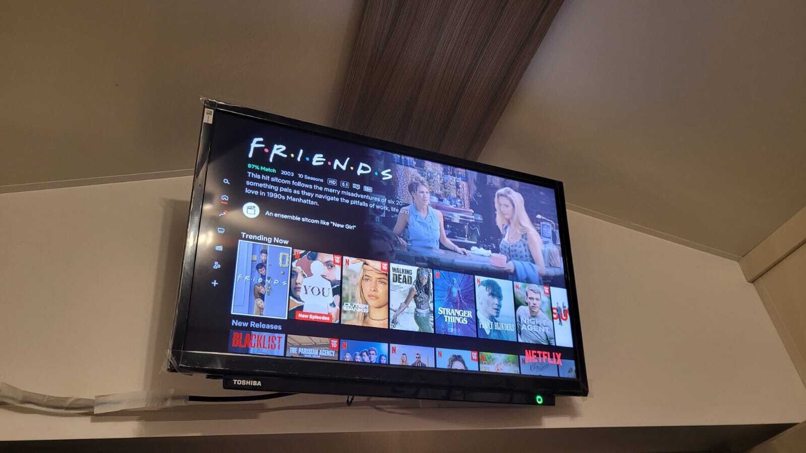 Un téléviseur à écran plat fixé au mur affiche l’émission « Friends », et l’interface Netflix apparaît en dessous.