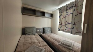 Un dormitorio pequeño y funcional con dos camas individuales, un estante empotrado sobre las camas y una ventana con una cortina colorida.