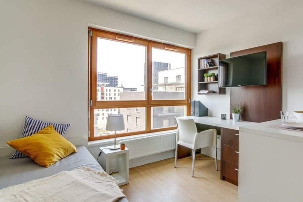 Ein Schlafzimmer mit Bett, Schreibtisch, Stuhl, Fernseher und Fenster mit Blick auf Gebäude.