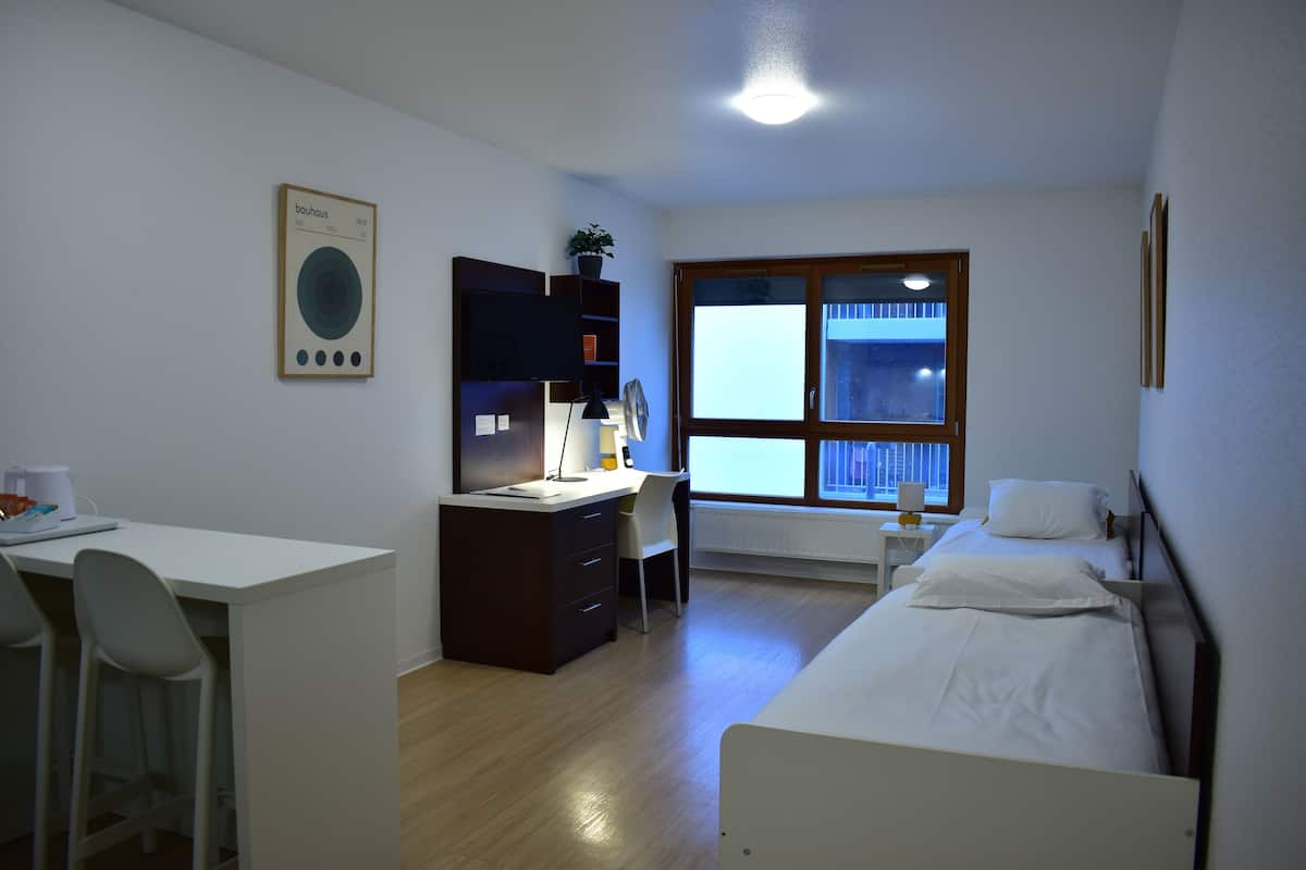 Ein kleiner, gut beleuchteter Raum mit Schreibtisch, Stuhl, Bett und Fenster mit Jalousien.
