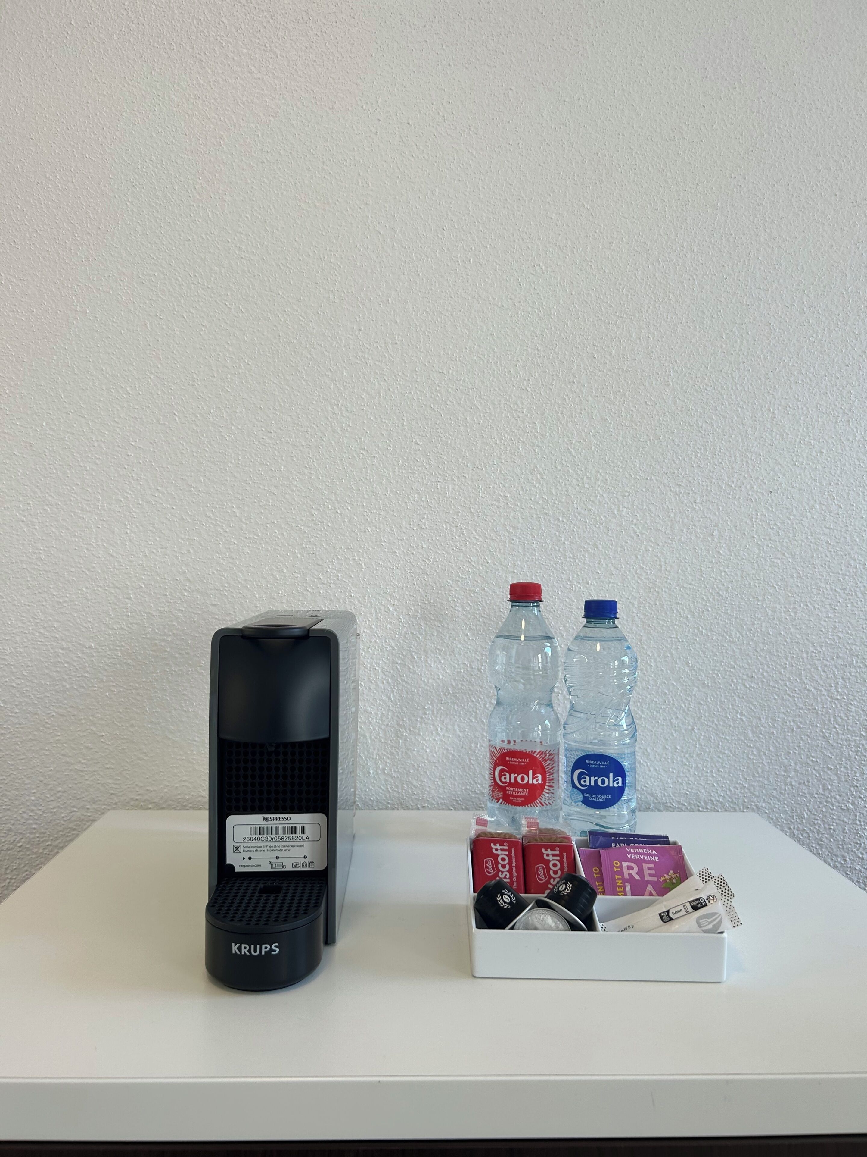 Une machine à café Krups, deux bouteilles d’eau et une boîte de capsules de café, le tout posé sur une surface blanche.