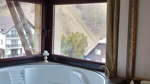 A bathroom with a freestanding bathtub, a window with curtains, a chandelier, and a view of buildings outside. - Vlahia Inn (Moieciu de Jos)