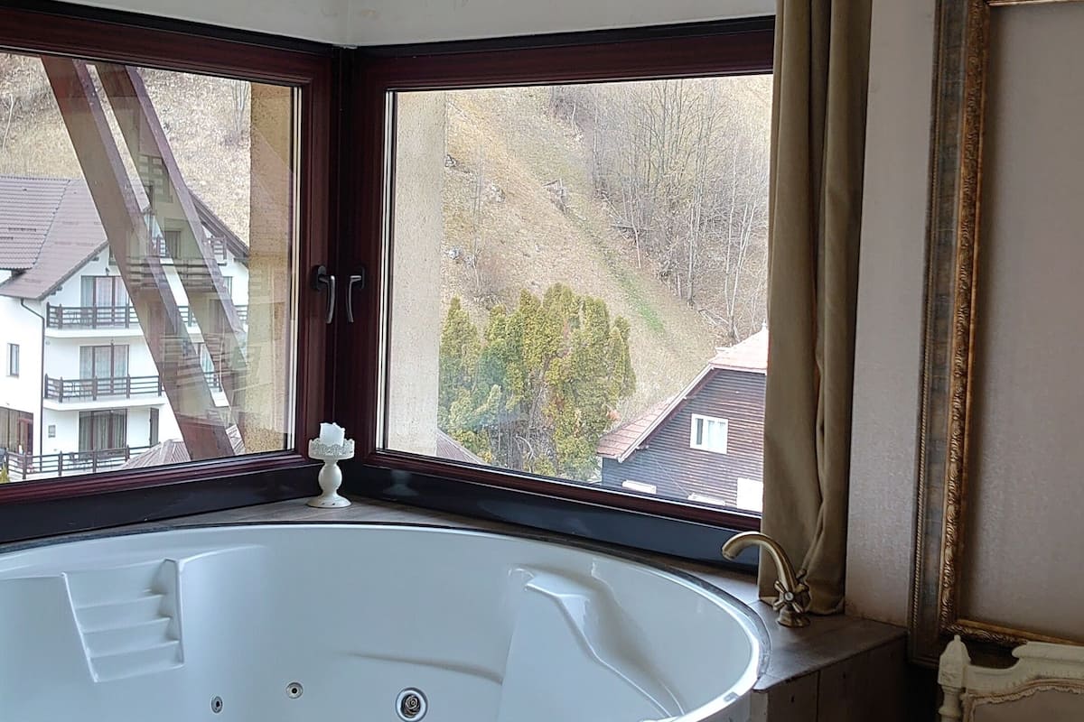 Ein Badezimmer mit einer freistehenden Badewanne, einem Fenster mit Vorhängen, einer Kristallleuchtre und Blick auf Gebäude.