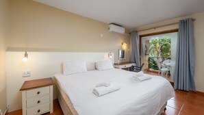 Un lit bien fait, avec du linge de lit et des serviettes blanches, une table de chevet avec des tiroirs, un petit bureau avec une chaise et un balcon donnant sur un espace verdoyant.