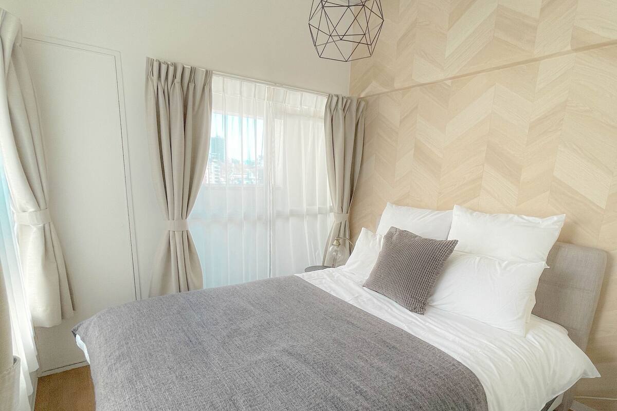 A bedroom with a large bed, a geometric pendant light, sheer curtains, and a herringbone-patterned wall.