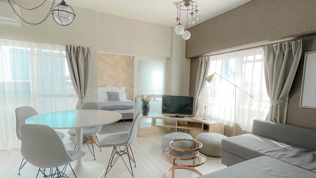 A modern living room with a large grey sofa, a dining table with chairs, a TV, and a chandelier. - Staypia Condominium Namba Ebisu (Osaka)