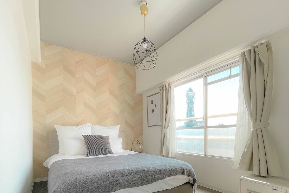 A modern bedroom with a bed, wooden floor, a geometric light fixture, a window with curtains, and a view of a building outside.