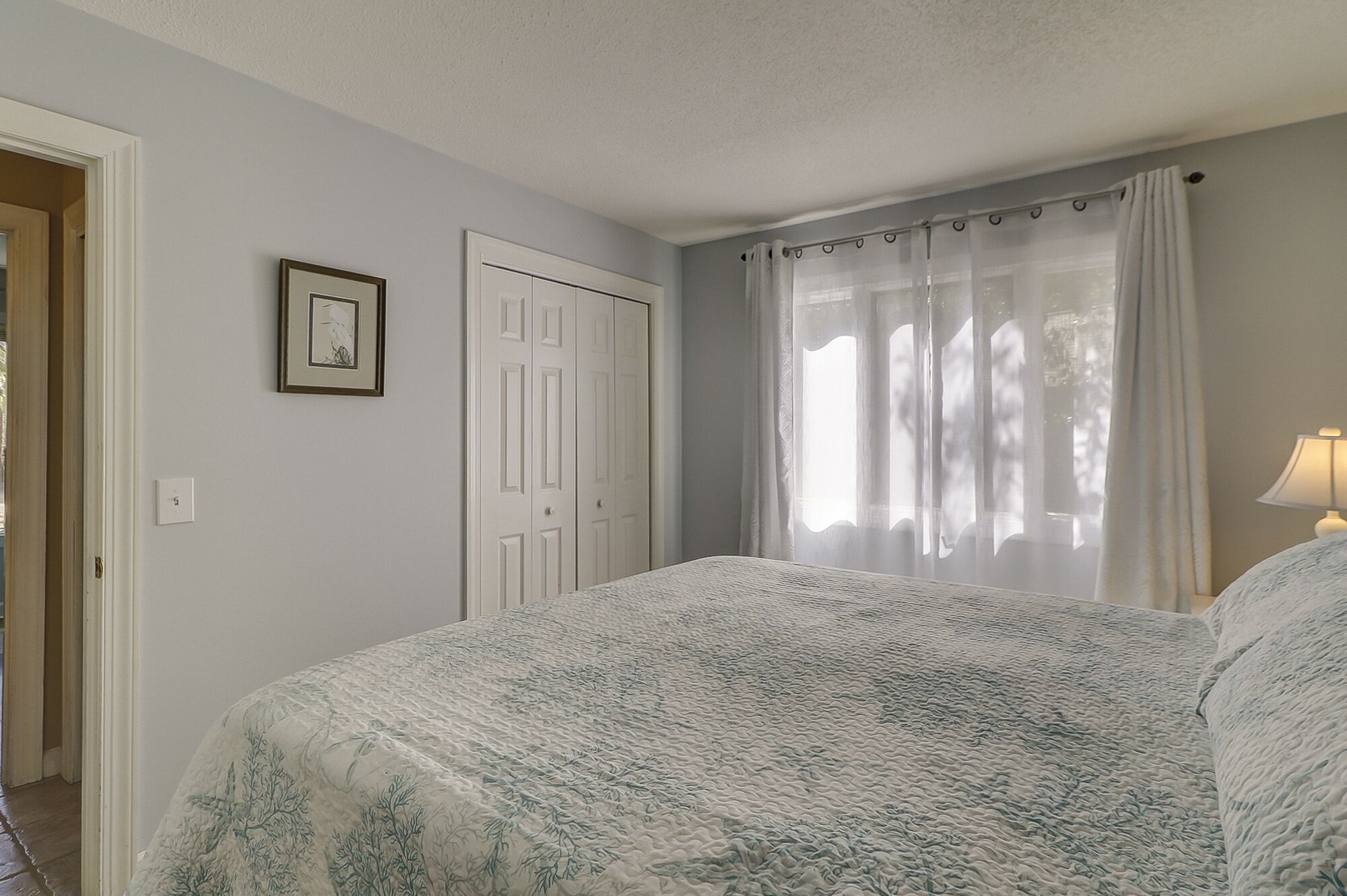 Un dormitorio con cama, una puerta, un cuadro enmarco y una ventana con cortinas.