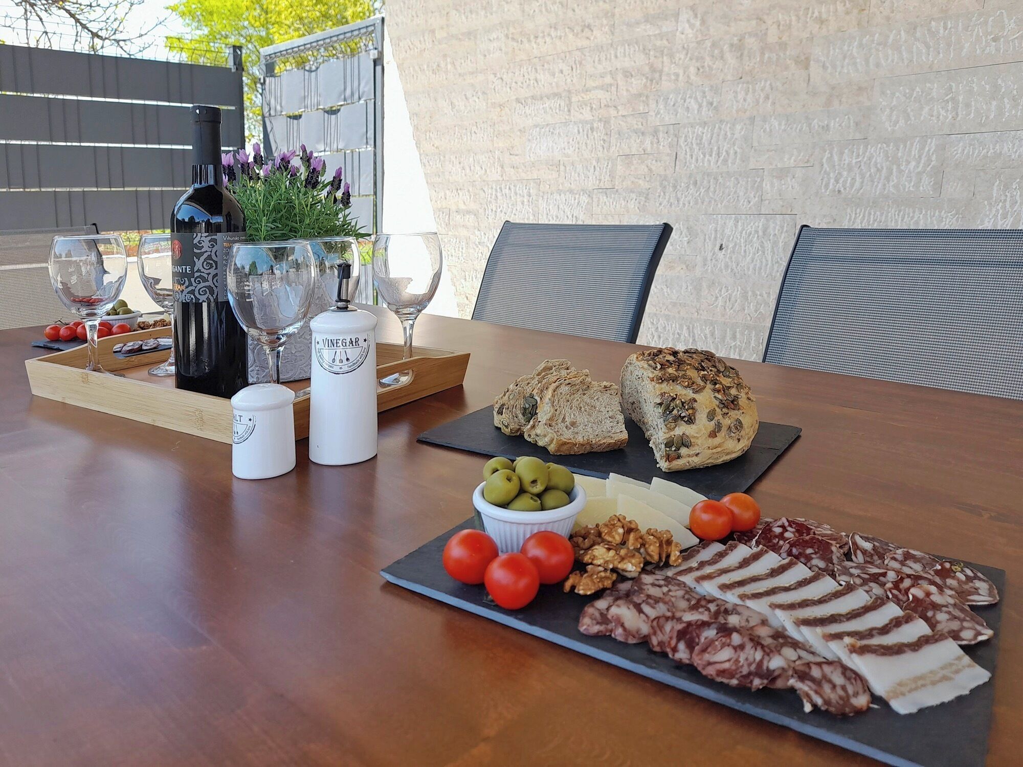 A wooden table with a bottle of wine, glasses, a bread basket, olives, tomatoes, walnuts, and various cheeses and cured meats.