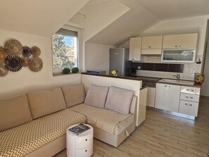 En moderne lejlighed med en opholdsstue med sofa og lille sofabord, et tekøkken med hvide skabe, samt et stort vindue der lukker naturligt lys ind.