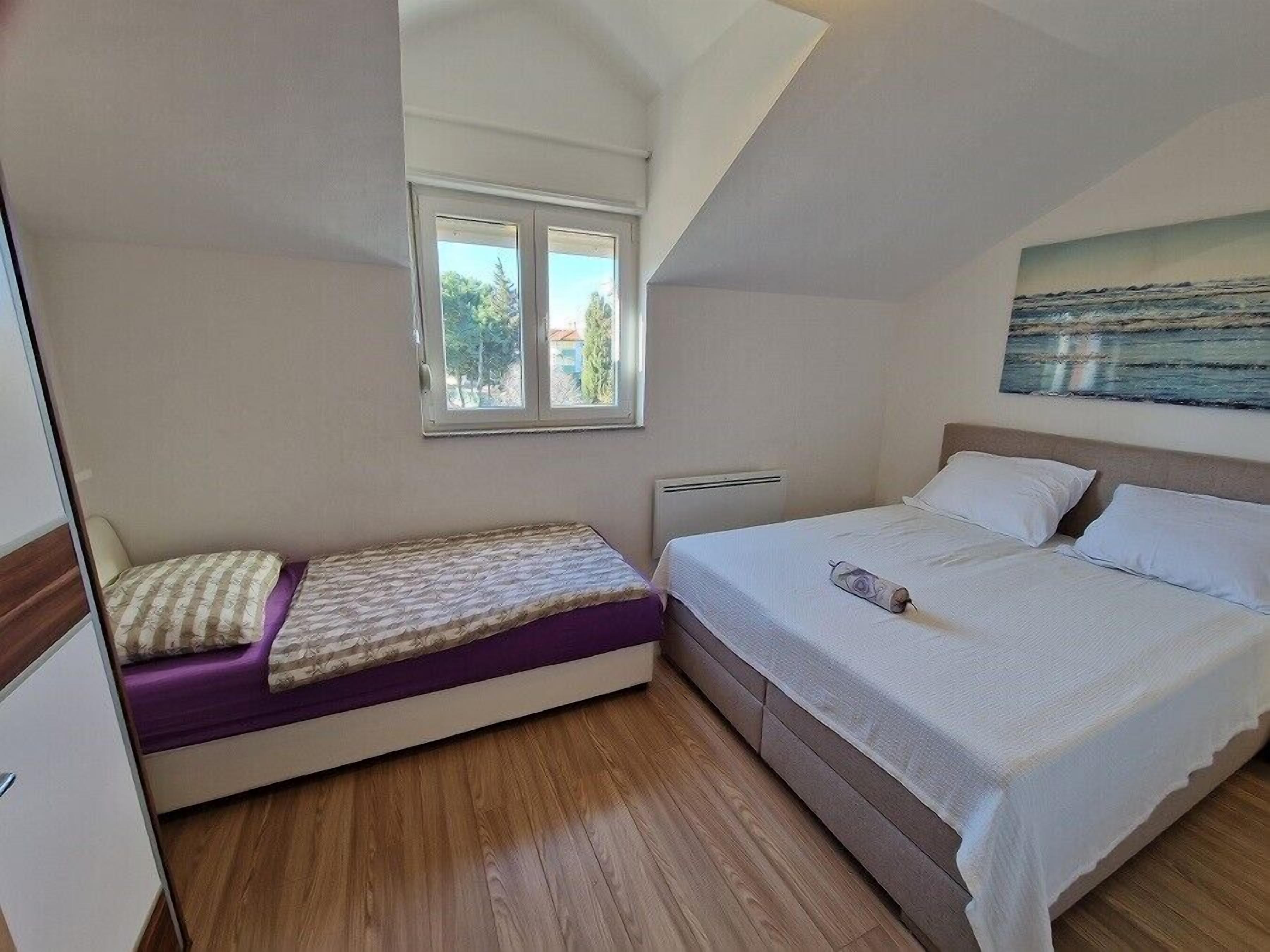 A bedroom with a single bed, a smaller bed, a window, and a painting on the wall.