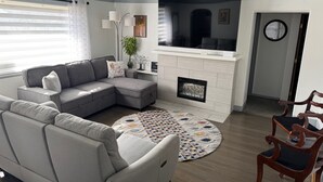 A living room with a sectional sofa, a fireplace, a television, and a dining area with a wooden chair. - Garland Charmer (Spokane)