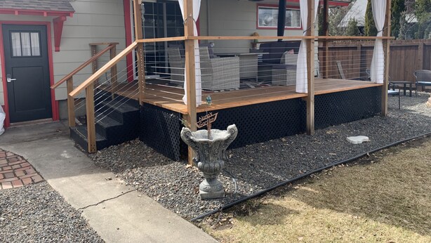 A covered outdoor deck with a wooden railing, a stone urn, and a sign reading "Naga's Garden." - Garland Charmer (Spokane)