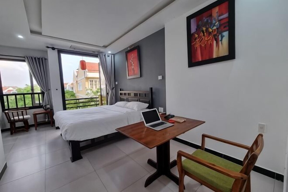 A hotel room with a bed, a desk with a laptop, a chair, and a balcony with a view.