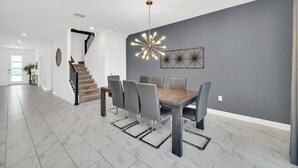 A modern dining area with a gray wall, a contemporary chandelier, and a staircase leading to another room. - 7 Bed Champions Gate 7 Bedroom Villa by RedAwning (Davenport)