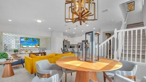 Una sala moderna con una mesa redonda de madera, sillas tapizadas de color gris, un sofá amarillo y un televisor montado en la pared.