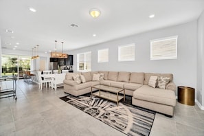 A modern living room with a large beige sectional sofa, a dining area with a white table and chairs, and a kitchen in the background. - 5 Bedroom Resort 5 bath Pool Home With Spa! 5 Villa by RedAwning (Davenport)