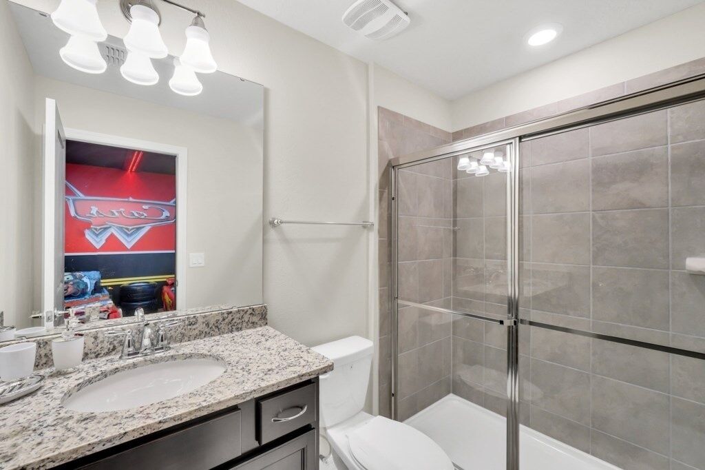 A bathroom with a granite countertop sink, a toilet, a glass shower enclosure, and a mirror with lights above.