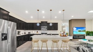 A modern kitchen with dark cabinets, stainless steel appliances, a central island with bar stools, and a flat-screen TV mounted on the wall. - Many Themes For Everyone 10 Huge Bedrooms! 10 Villa by RedAwning (Kissimmee)