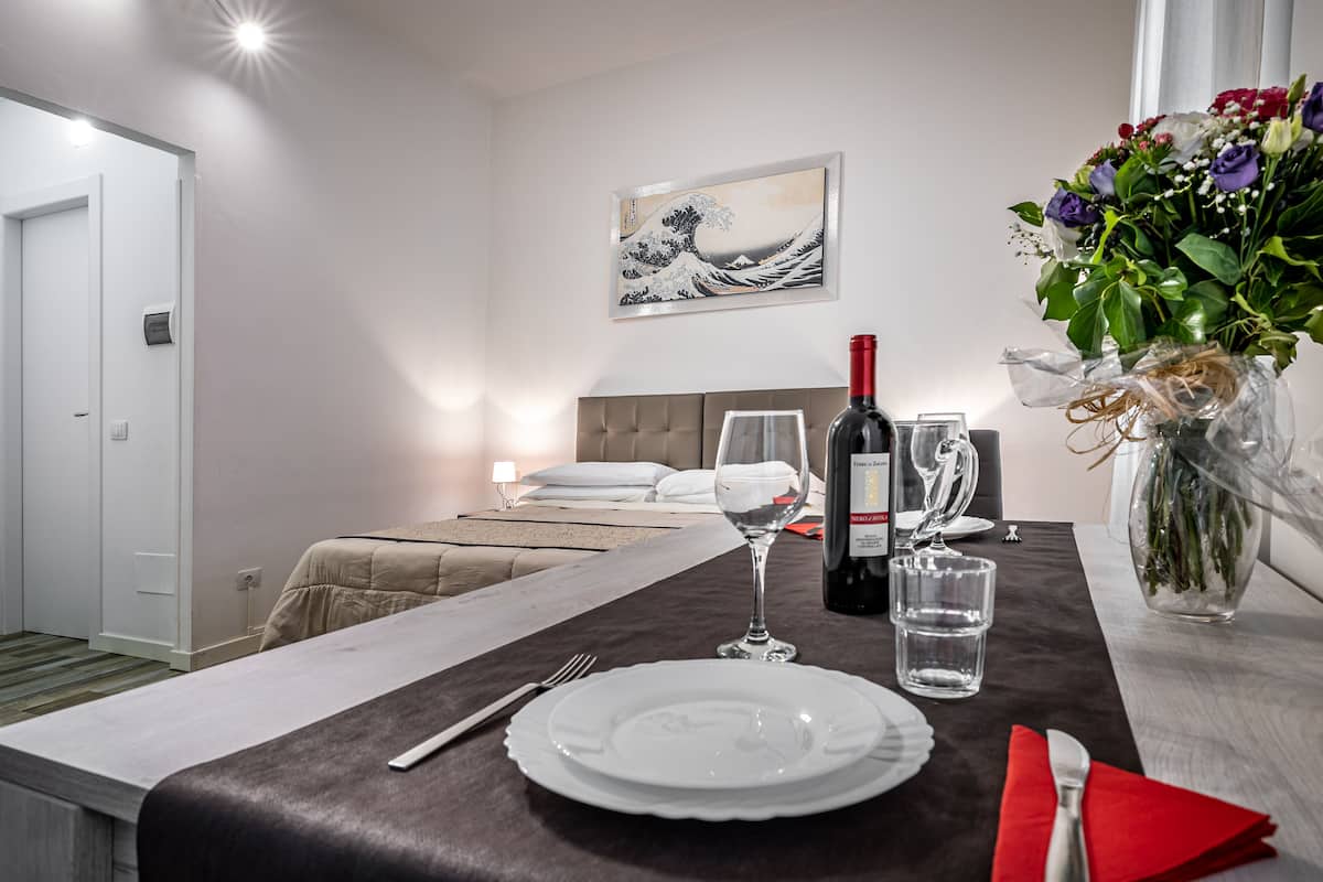 A hotel room with a neatly made bed, a dining table set with plates, glasses, and a bottle of wine, and a vase of flowers on the table.