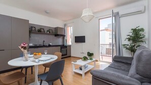 En moderne stue med en grå sofa, et spisebord med stole, et lille hvidt sofabord og et køkkenområde med et fjernsyn.