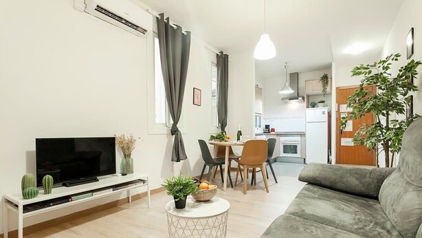 En moderne stue med et fladskærms-tv, en grå sofa, et spiseområde med bord og stole, og et køkken i baggrunden.