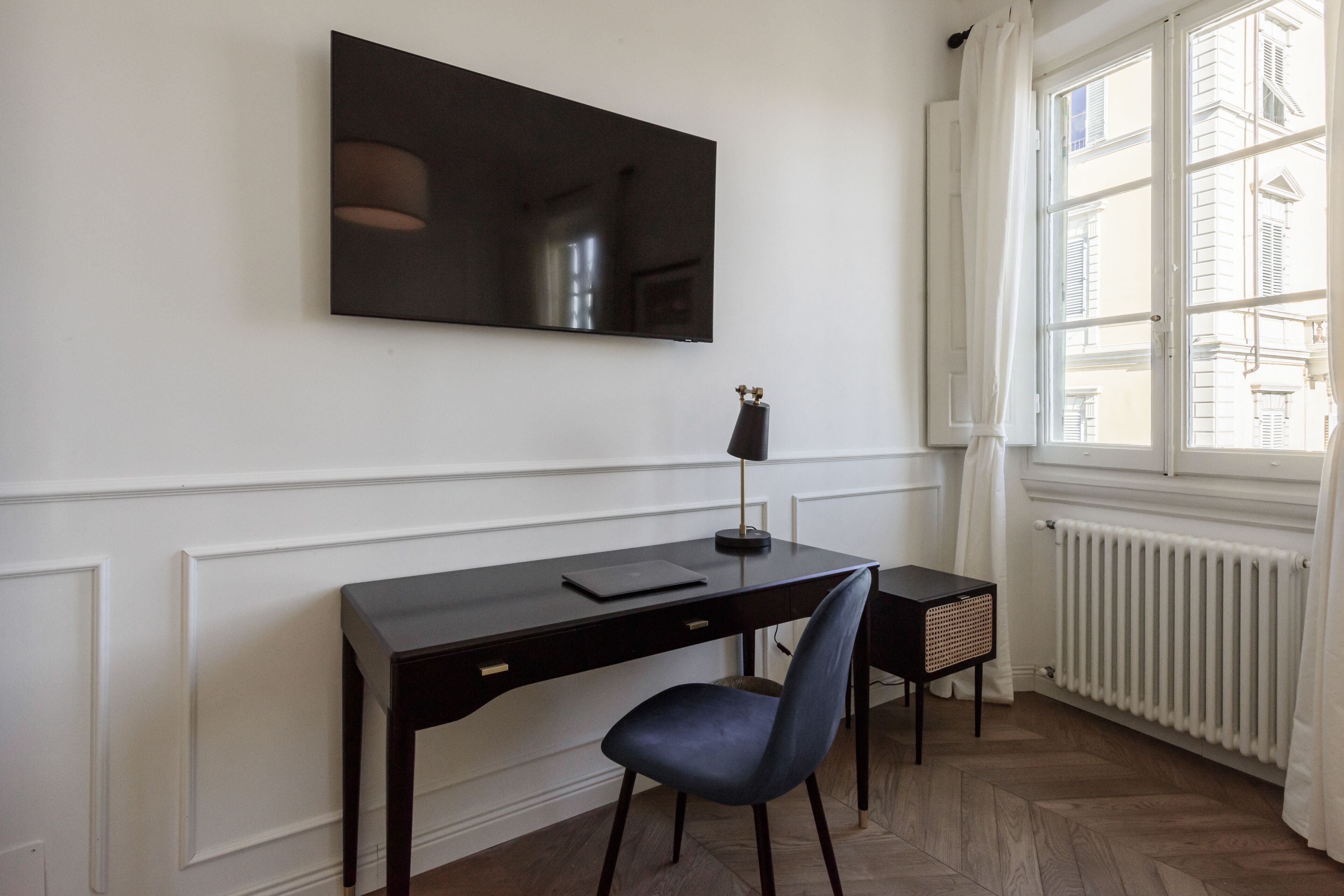 A room with a dark wooden desk, a black chair, a flat-screen TV mounted on the wall, a small side table with a lamp, and a window with white curtains.