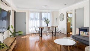 A modern living room with a wooden floor, a dining area with a table and chairs, a grey sofa, and a large window with sheer curtains.