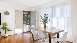 A dining area with a wooden table, white chairs, a potted plant, and a clock on the wall.