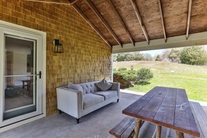 An outdoor patio with a wooden table and a sofa, a brick wall, and a glass door leading to an interior space.
