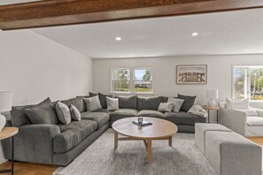 Un salon moderne avec un canapé d’angle, une table basse ronde et une vue sur l’extérieur grâce à de grandes fenêtres.